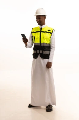 استخدام التكنولوجيا والتقنيات الحديثة فى العمل ،المتابعة والإشراف على العمل عبر الجوال، الإعمار بسواعد أبناء الوطن، مفهوم الهندسة والانشاءات المعمارية، بورتريه لمهندس عربي خليجي سعودي اسمر يرتدي الثوب التقليدي والسترة الخاصة وخوذة الحماية يقف وبيده الهاتف المحمول ، الجسم بالطول الكامل، خلفية بيضاء