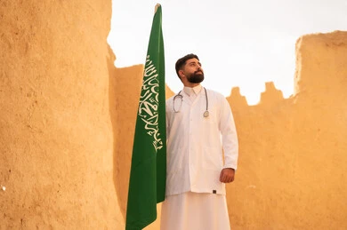 الإحتفال بيوم العلم 11 مارس، مهن ووظائف سعودية طبية، مفهوم العمل في القطاع الطبي، طبيب عربي خليجي سعودي يرتدي الثوب التقليدي المعطف الطبي والسماعات يقف في بيت طيني ويحمل بيده علم المملكة، إحياء ذكرى اليوم الوطني السعودي 23 سبتمبر، يوم بدينا 1727 م