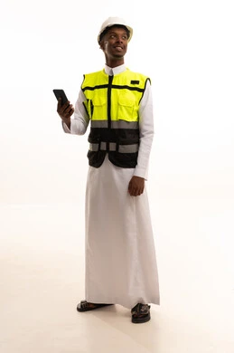 مفهوم الهندسة والانشاءات المعمارية، المتابعة والإشراف على العمل عبر الجوال، الإعمار بسواعد أبناء الوطن، استخدام التكنولوجيا والتقنيات الحديثة فى العمل ، بورتريه لمهندس عربي خليجي سعودي اسمر يرتدي الثوب التقليدي والسترة الخاصة وخوذة الحماية يقف وبيده الهاتف المحمول ، الجسم بالطول الكامل، خلفية بيضاء