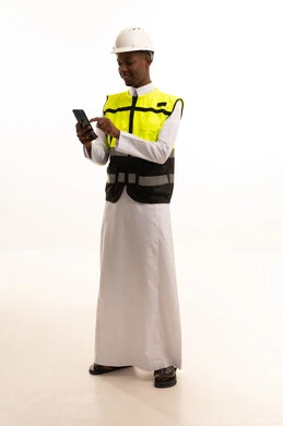 الإعمار بسواعد أبناء الوطن، مفهوم الهندسة والانشاءات المعمارية، استخدام التكنولوجيا والتقنيات الحديثة فى العمل ،المتابعة والإشراف على العمل عبر الجوال،بورتريه لمهندس عربي خليجي سعودي اسمر يرتدي الثوب التقليدي والسترة الخاصة وخوذة الحماية يقف وبيده الهاتف المحمول ، الجسم بالطول الكامل، خلفية بيضاء