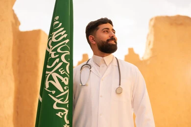 مهن ووظائف سعودية طبية، مفهوم العمل في القطاع الطبي، طبيب عربي خليجي سعودي يرتدي الثوب التقليدي المعطف الطبي والسماعات يقف في بيت طيني ويحمل بيده علم المملكة، إحياء ذكرى اليوم الوطني السعودي 23 سبتمبر، يوم بدينا 1727 م، الإحتفال بيوم العلم 11 مارس