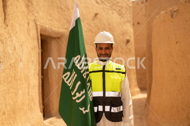 رفع علم المملكة في الأماكن التراثيه الشعبيه في 11 مارس، فن العمارة القديم وبناء البيوت الطينية، مهندس عربي خليجي سعودي مبتسم يرتدي السترة وخوذة الحماية بايماءات الفخر والاعتزاز في اليوم الوطني 23 سبتمبر، يوم بدينا 1727 م، ذكرى تأسيس الدولة السعودية الأولى 22 فبراير