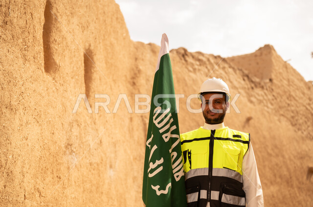 Raising the Kingdom's flag in popular heritage sites on March 11, ancient architecture and mud house construction, a smiling Saudi Gulf Arab engineer wearing a jacket and protective helmet with gestures of pride and honor on National Day, September 23, the day we began in 1727 AD, the anniversary of the founding of the first Saudi state, February 22