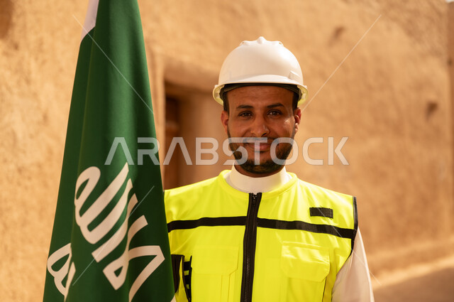Raising the Kingdom's flag in popular heritage sites on March 11, ancient architecture and mud house construction, a smiling Saudi Gulf Arab engineer wearing a jacket and protective helmet with gestures of pride and honor on National Day, September 23, the day we began in 1727 AD, the anniversary of the founding of the first Saudi state, February 22