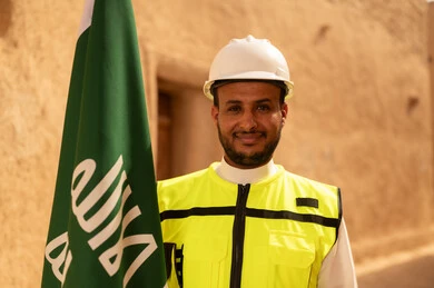 Raising the Kingdom's flag in popular heritage sites on March 11, ancient architecture and mud house construction, a smiling Saudi Gulf Arab engineer wearing a jacket and protective helmet with gestures of pride and honor on National Day, September 23, the day we began in 1727 AD, the anniversary of the founding of the first Saudi state, February 22