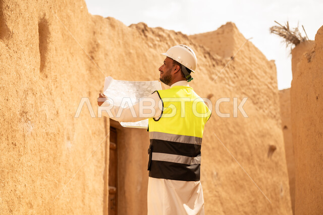 اساسيات البناء للبيوت والقصور التراثية، يوم بدينا 1727 م، اليوم الوطني 23 سبتمبر، التعرف على فن العمارة القديمة، ذكرى تأسيس الدولة السعودية الأولى 22 فبراير، مهندس عربي خليجي سعودي يرتدي السترة والخوذة يمسك بيده مخطط البناء، التجول في الحي الشعبي
