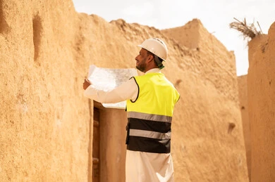 اساسيات البناء للبيوت والقصور التراثية، يوم بدينا 1727 م، اليوم الوطني 23 سبتمبر، التعرف على فن العمارة القديمة، ذكرى تأسيس الدولة السعودية الأولى 22 فبراير، مهندس عربي خليجي سعودي يرتدي السترة والخوذة يمسك بيده مخطط البناء، التجول في الحي الشعبي