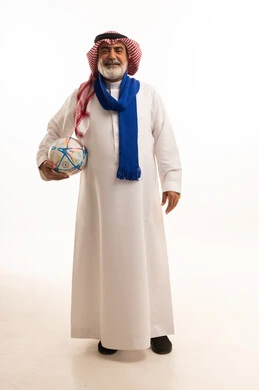 Enthusiasm and interaction with the national team, passion for supporting different sports clubs, portrait of an elderly Saudi Arabian Gulf man wearing a traditional shemagh and thobe, wearing a scarf and holding a football, full body, white background