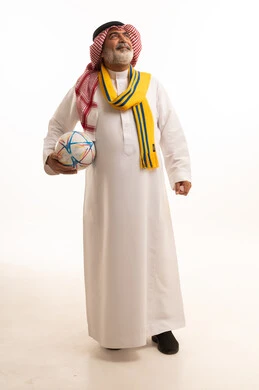 الشغف في تشجيع الأنديه الرياضية المختلفة، الحماس والتفاعل مع المنتخب، بورتريه لرجل كبير السن عربي خليجي سعودي يرتدي الشماغ والثوب التقليدي يضع الوشاح يحمل بيده كرة القدم، الجسم بالطول الكامل، خلفية بيضاء