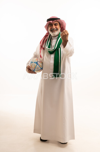 Winning, profit and triumph, enthusiasm and interaction with the team, portrait of an elderly Saudi Arabian Gulf man wearing a traditional shemagh and thobe, wearing a scarf and holding a football, different sports clubs, full body, white background