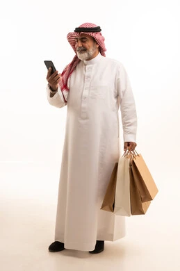 شراء المنتجات و الأغراض عبر الانترنت, بورتريه لرجل عربي خليجي سعودي كبير في السن يرتدي الثوب التقليدي و الشماغ يمسك بيده أكياس ورقية والهاتف المحمول, صورة للجسم بالطول الكامل , خلفية بيضاء