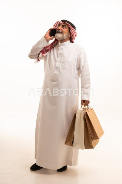 اجراء مكالمة هاتفية عبر الجوال، شراء المنتجات و الأغراض عبر الانترنت, بورتريه لرجل عربي خليجي سعودي كبير في السن يرتدي الثوب التقليدي و الشماغ يمسك بيده أكياس ورقية والهاتف المحمول, صورة للجسم بالطول الكامل , خلفية بيضاء