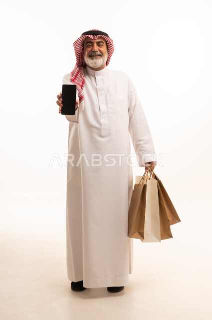 View blank black screen on mobile, buy products and items online, portrait of elderly Saudi Arabian Gulf man wearing traditional thobe and shemagh holding paper bags and mobile phone, full body photo, white background