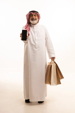 View blank black screen on mobile, buy products and items online, portrait of elderly Saudi Arabian Gulf man wearing traditional thobe and shemagh holding paper bags and mobile phone, full body photo, white background