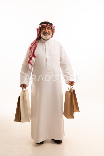 الاستفادة من الحسومات الموسمية، إيماءات السعادة والسرور بالخصومات، بورتريه لرجل كبير بالسن عربي خليجي سعودي يرتدي الشماغ و الثوب التقليدي يحمل بيده الاكياس الورقية، التسوق والحصول على العروض، صورة الجسم بالطول الكامل، خلفية بيضاء