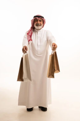 الاستفادة من الحسومات الموسمية، إيماءات السعادة والسرور بالخصومات، بورتريه لرجل كبير بالسن عربي خليجي سعودي يرتدي الشماغ و الثوب التقليدي يحمل بيده الاكياس الورقية، التسوق والحصول على العروض، صورة الجسم بالطول الكامل، خلفية بيضاء