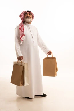 Taking advantage of seasonal discounts, gestures of happiness and pleasure with discounts, portrait of an elderly Saudi Arabian Gulf man wearing a traditional shemagh and thobe holding paper bags, shopping and getting offers, full body image, white background