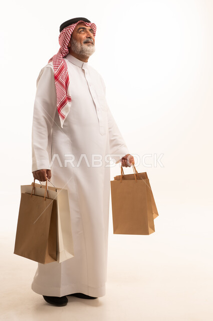 Taking advantage of seasonal discounts, gestures of happiness and pleasure with discounts, portrait of an elderly Saudi Arabian Gulf man wearing a traditional shemagh and thobe holding paper bags, shopping and getting offers, full body image, white background