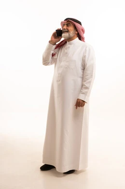 اجراء مكالمة هاتفية عبر الجوال، تطور التكنولوجيا والتقنيات الحديثة، بورتريه لرجل كبير السن عربي سعودي خليجي يرتدي الشماغ والثوب التقليدي يقوم باستخدام الهاتف المحمول، صورة الجسم بالطول الكامل، خلفية بيضاء