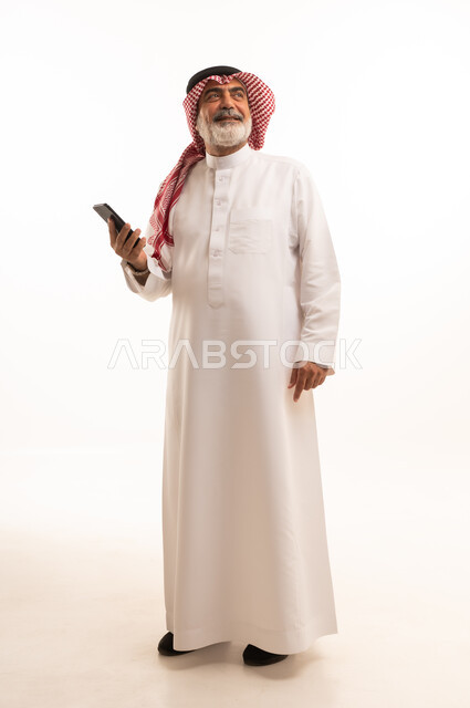 تطور التكنولوجيا والتقنيات الحديثة، تصفح مواقع التواصل الاجتماعي عبر الانترنت، بورتريه لرجل كبير السن عربي سعودي خليجي يرتدي الشماغ والثوب التقليدي يقوم باستخدام الهاتف المحمول، صورة الجسم بالطول الكامل، خلفية بيضاء