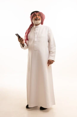 تطور التكنولوجيا والتقنيات الحديثة، تصفح مواقع التواصل الاجتماعي عبر الانترنت، بورتريه لرجل كبير السن عربي سعودي خليجي يرتدي الشماغ والثوب التقليدي يقوم باستخدام الهاتف المحمول، صورة الجسم بالطول الكامل، خلفية بيضاء