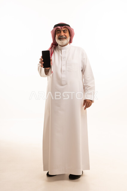 Blank black screen view on mobile, development of technology and modern techniques, portrait of elderly Saudi Arabian Gulf man wearing traditional shemagh and thobe using mobile phone, full body image, white background