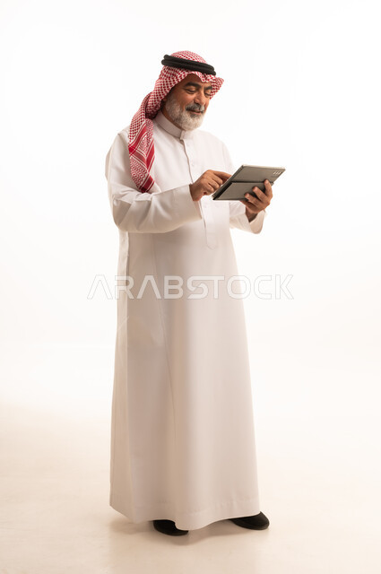دمج التقنية في الحياة اليومية، بورتريه لرجل عربي خليجي سعودي كبير بالسن يرتدي الشماغ والثوب التقليدي يعمل على جهاز التابلت، تطور التكنولوجيا والأجهزة التقنية، استخدام الجهاز اللوحي والعمل عن بعد، صورة الجسم بالطول الكامل، خلفية بيضاء