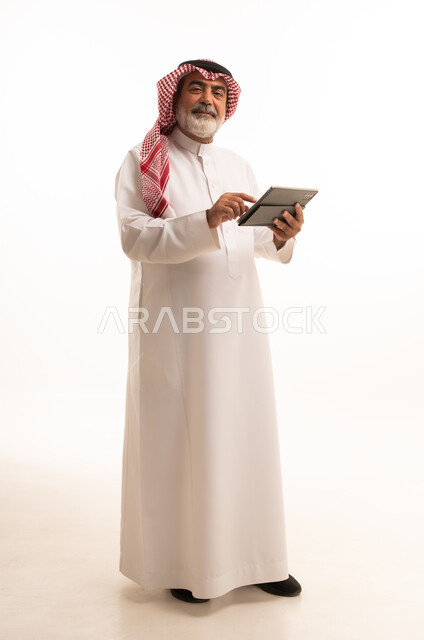 دمج التقنية في الحياة اليومية، بورتريه لرجل عربي خليجي سعودي كبير بالسن يرتدي الشماغ والثوب التقليدي يعمل على جهاز التابلت، تطور التكنولوجيا والأجهزة التقنية، استخدام الجهاز اللوحي والعمل عن بعد، صورة الجسم بالطول الكامل، خلفية بيضاء