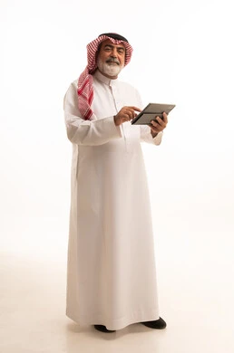 دمج التقنية في الحياة اليومية، بورتريه لرجل عربي خليجي سعودي كبير بالسن يرتدي الشماغ والثوب التقليدي يعمل على جهاز التابلت، تطور التكنولوجيا والأجهزة التقنية، استخدام الجهاز اللوحي والعمل عن بعد، صورة الجسم بالطول الكامل، خلفية بيضاء