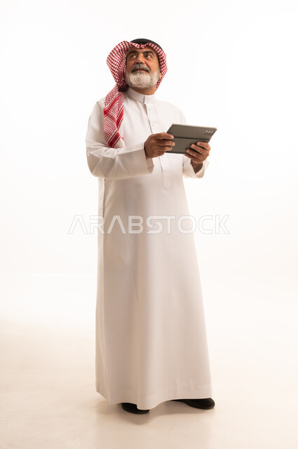 دمج التقنية في الحياة اليومية، بورتريه لرجل عربي خليجي سعودي كبير بالسن يرتدي الشماغ والثوب التقليدي يعمل على جهاز التابلت، تطور التكنولوجيا والأجهزة التقنية، استخدام الجهاز اللوحي والعمل عن بعد، صورة الجسم بالطول الكامل، خلفية بيضاء