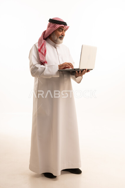 استخدام اللابتوب والعمل عن بعد، دمج التقنية في الحياة اليومية، بورتريه لرجل عربي خليجي سعودي كبير بالسن يرتدي الشماغ والثوب التقليدي يحمل الحاسوب المحمول، تطور التكنولوجيا والأجهزة التقنية، صورة الجسم بالطول الكامل، خلفية بيضاء