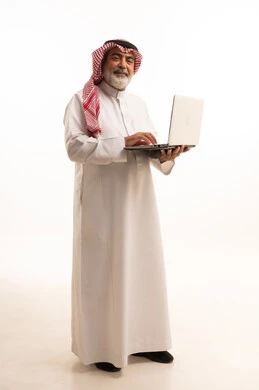 استخدام اللابتوب والعمل عن بعد، دمج التقنية في الحياة اليومية، بورتريه لرجل عربي خليجي سعودي كبير بالسن يرتدي الشماغ والثوب التقليدي يحمل الحاسوب المحمول، تطور التكنولوجيا والأجهزة التقنية، صورة الجسم بالطول الكامل، خلفية بيضاء