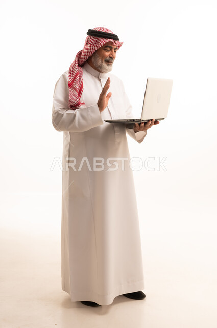 استخدام اللابتوب والعمل عن بعد، دمج التقنية في الحياة اليومية، بورتريه لرجل عربي خليجي سعودي كبير بالسن يرتدي الشماغ والثوب التقليدي يحمل الحاسوب المحمول، تطور التكنولوجيا والأجهزة التقنية، صورة الجسم بالطول الكامل، خلفية بيضاء
