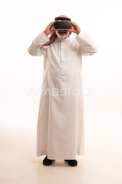 قضاء أوقات ممتعة ومسلية بالألعاب الافتراضية، الاستمتاع بمشاهدة أفلام الواقع الافتراضي، بورتريه لرجل عربي خليجي سعودي كبير بالسن يرتدي الثوب التقليدي و الشماغ يضع نظارة الواقع الافتراضي ثلاثية الابعاد، الجسم بالطول الكامل، خلفية بيضاء