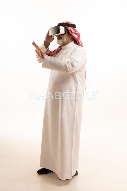 قضاء أوقات ممتعة ومسلية بالألعاب الافتراضية، الاستمتاع بمشاهدة أفلام الواقع الافتراضي، بورتريه لرجل عربي خليجي سعودي كبير بالسن يرتدي الثوب التقليدي و الشماغ يضع نظارة الواقع الافتراضي ثلاثية الابعاد، الجسم بالطول الكامل، خلفية بيضاء