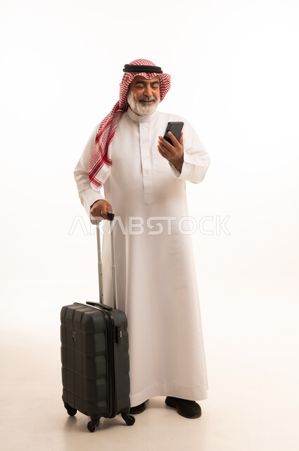 استخدام الأجهزة الحديثة و المتطورة , التجهيز للسفر والرحلات الممتعة, بورتريه لرجل عربي خليجي سعودي كبير في السن يرتدي الثوب التقليدي والشماغ يمسك بيده حقيبة السفر والهاتف المحمول, صورة للجسم بالطول الكامل , خلفية بيضاء