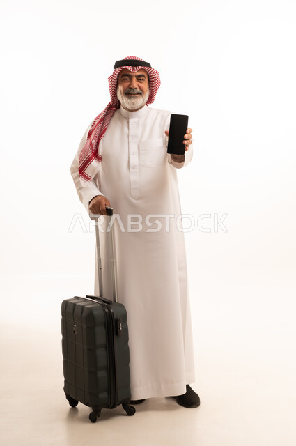 Empty black screen, using modern and advanced devices, preparing for travel and enjoyable trips, portrait of an elderly Saudi Arabian Gulf man wearing traditional thobe and shemagh holding a travel bag and mobile phone, full-length body image, white background