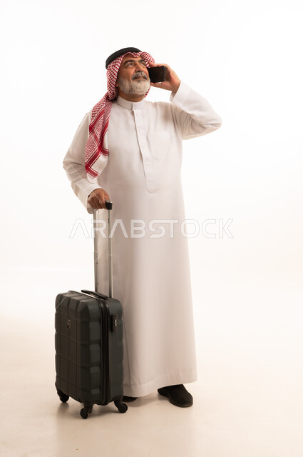 اجراء مكالمة هاتفية، استخدام الأجهزة الحديثة و المتطورة , التجهيز للسفر والرحلات الممتعة, بورتريه لرجل عربي خليجي سعودي كبير في السن يرتدي الثوب التقليدي والشماغ يمسك بيده حقيبة السفر والهاتف المحمول, صورة للجسم بالطول الكامل , خلفية بيضاء