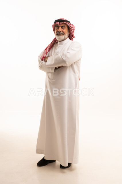 التقدم بالعمر والهيبة، بورتريه لرجل كبير بالسن عربي خليجي سعودي يرتدي الثوب التقليدي و الشماغ يقف بأيدي متقاطعة بتعابير الثقة بالنفس, الاهتمام بالأناقة والمظهر الخارجي، مفهوم الرجولة العربية, الجسم بالطول الكامل، خلفية بيضاء