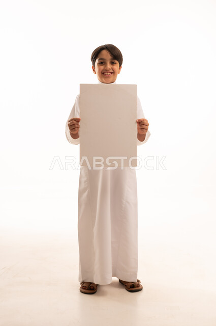 Looking at camera with pleased gestures, white square mockup, blank billboard, use of illustrative panels, portrait of smiling Saudi Arabian Gulf boy wearing traditional thobe standing straight and holding a white square panel, full body, white background