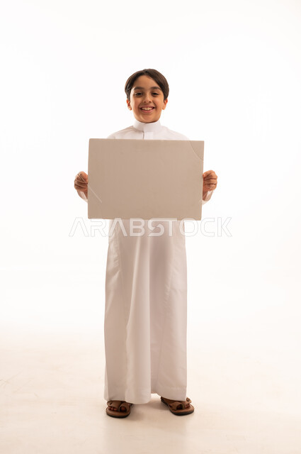 Looking at camera with pleased gestures, white square mockup, blank billboard, use of illustrative panels, portrait of smiling Saudi Arabian Gulf boy wearing traditional thobe standing straight and holding a white square panel, full body, white background