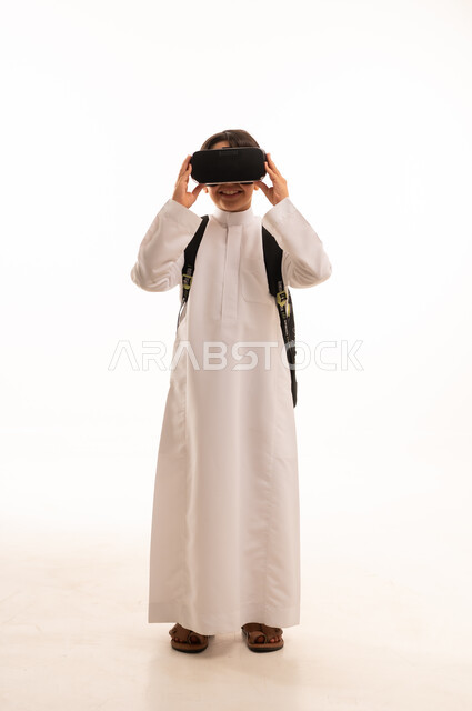 نشاطات الواقع الافتراضي، الدراسة عبر المنصات تعليمية، محاكاة العالم الخيالي، بورتريه لطالب عربي خليجي سعودي يرتدي الثوب التقليدي وحقيبة الظهر المدرسية يضع نظارة الفي آر، صورة الجسم بالكامل، خلفية بيضاء