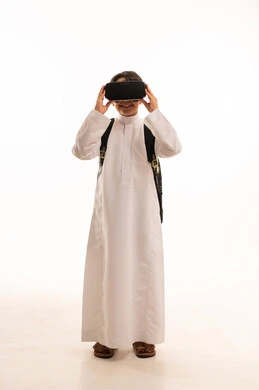 نشاطات الواقع الافتراضي، الدراسة عبر المنصات تعليمية، محاكاة العالم الخيالي، بورتريه لطالب عربي خليجي سعودي يرتدي الثوب التقليدي وحقيبة الظهر المدرسية يضع نظارة الفي آر، صورة الجسم بالكامل، خلفية بيضاء