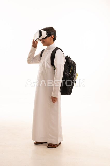 الدراسة عبر المنصات تعليمية، محاكاة العالم الخيالي، بورتريه لطالب عربي خليجي سعودي يرتدي الثوب التقليدي وحقيبة الظهر المدرسية يضع نظارة الفي آر، نشاطات الواقع الافتراضي، صورة الجسم بالكامل، خلفية بيضاء