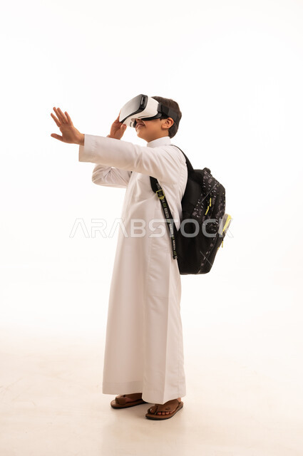 نشاطات الواقع الافتراضي، الدراسة عبر المنصات تعليمية، محاكاة العالم الخيالي، بورتريه لطالب عربي خليجي سعودي يرتدي الثوب التقليدي وحقيبة الظهر المدرسية يضع نظارة الفي آر، صورة الجسم بالكامل، خلفية بيضاء