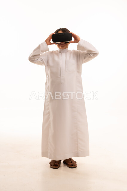 محاكاة العالم الخيالي والنظر الى الافلام الواقعيه، تعابير الاندماج والتركيز، التطور التقني والتكنولوجي، بورتريه لولد عربي خليجي سعودي يرتدي الثوب التقليدي ينظر من خلال نظارات ثلاثية الأبعاد (الفي ار),  الجسم بالطول الكامل، خلفيه بيضاء