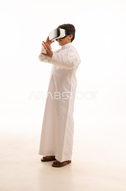 Simulating the imaginary world and looking at real movies, expressions of integration and concentration, technical and technological development, portrait of a Saudi Arabian Gulf boy wearing traditional dress looking through 3D glasses (VR), full body, white background