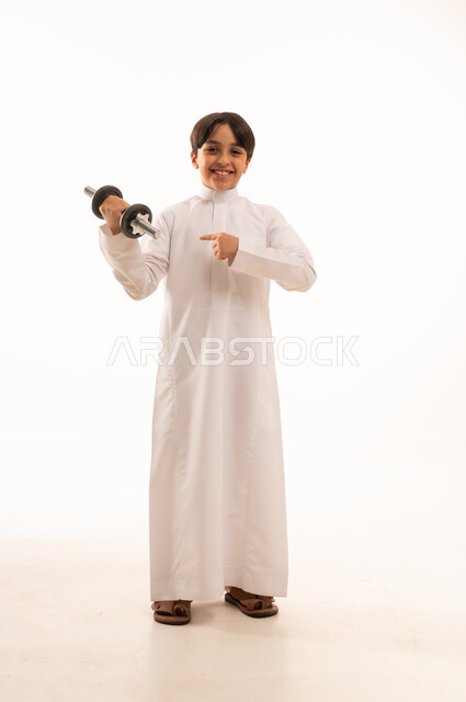 هواية ممارسة الرياضة, بورتريه لولد عربي خليجي يرتدي الثوب التقليدي الأبيض يحمل بيده الأوزان الثقيلة الحديدية ( الدمبلز) , النظر للكاميرا بتعابير القوة, أنشطة رياضية ترفيهية للأولاد، الجسم بالطول الكامل، خلفية بيضاء