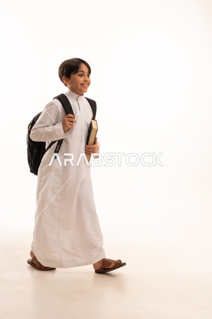 مفهوم الاجتهاد للوصول إلى النجاح، المناهج العلمية التعليمية، مفهوم العودة إلى المدارس، بورتريه لطالب عربي خليجي سعودي يرتدي الثوب التقليدي يحمل حقيبة الظهر المدرسية ويحمل بيده كتاب، صورة لكامل الجسم، خلفية بيضاء