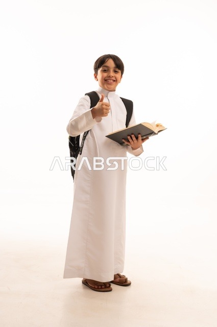 رفع اصبع الابهام بايماءات الرضى والقبول، الاجتهاد للوصول إلى النجاح، المناهج العلمية التعليمية، مفهوم العودة إلى المدارس، بورتريه لطالب عربي خليجي سعودي يرتدي الثوب التقليدي يحمل حقيبة الظهر المدرسية ويقوم بقراءة كتاب، صورة لكامل الجسم، خلفية بيضاء